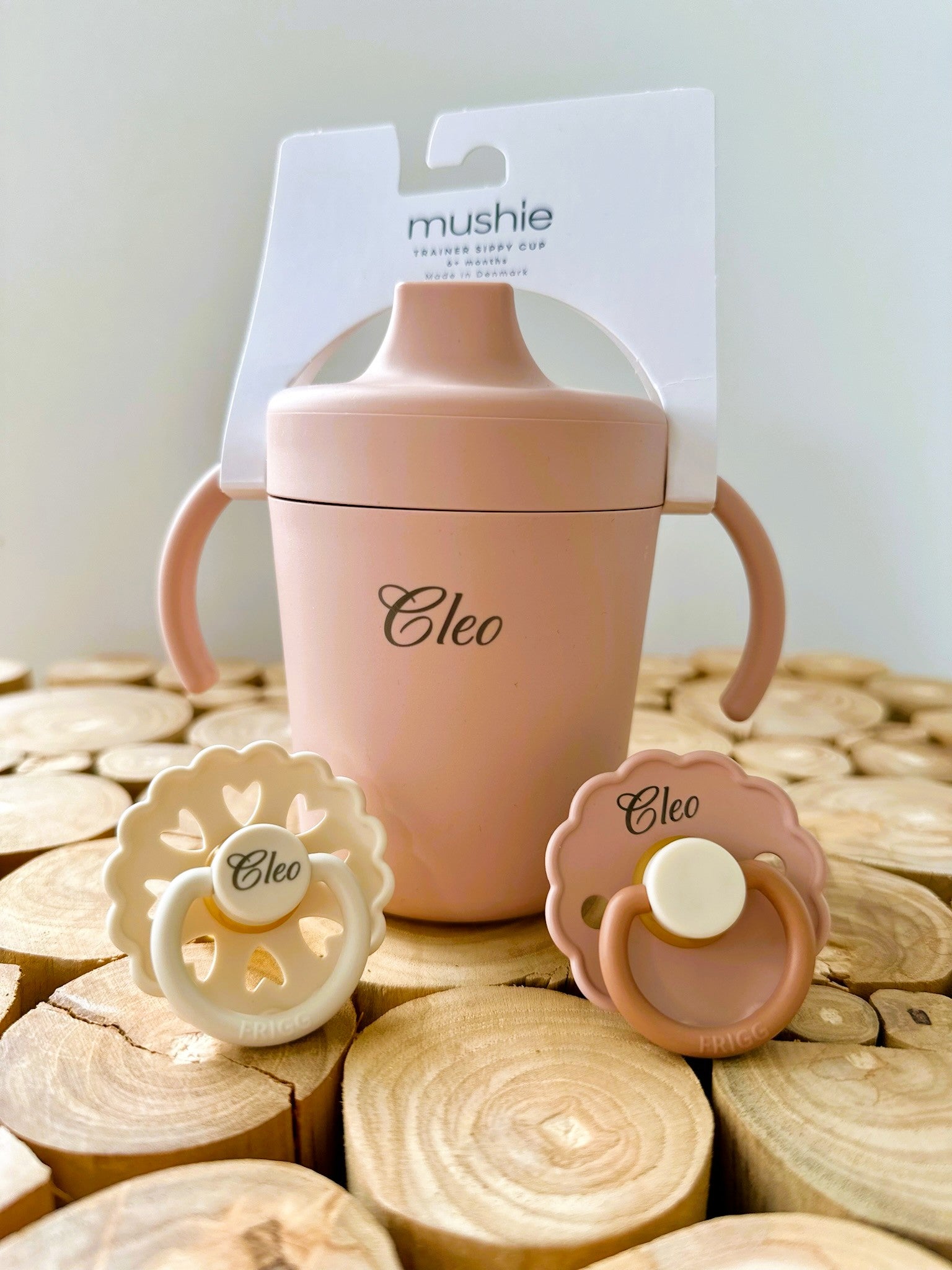 Gepersonaliseerde Mushie drinkbeker en twee Frigg spenen met de naam "Cleo", in zachte beige en roze tinten, gepresenteerd op een houten achtergrond.